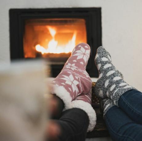 pogled od zadaj na dve prijateljici, ki sedita ob kaminu in božično drevo z nogami z zimskimi nogavicami na mizici, pijeta kavo ali čaj